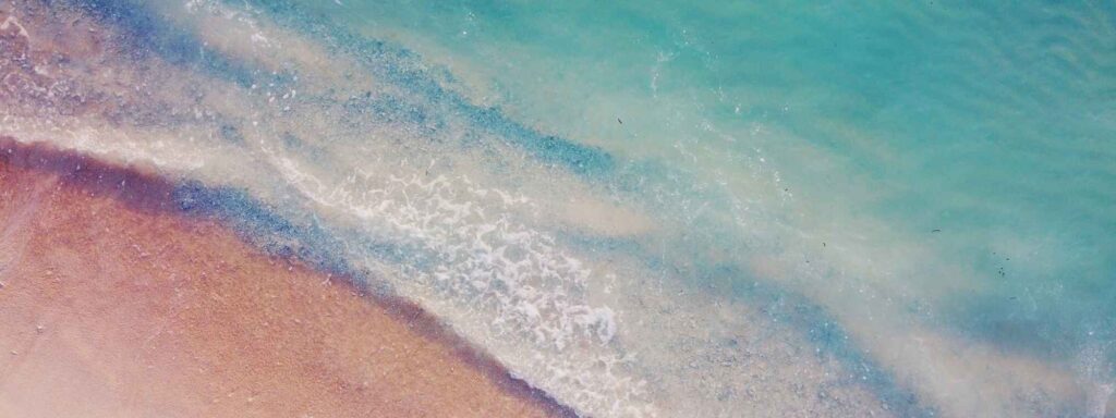 Aerial view of pink sand meeting turquoise ocean with foamy waves arriving on shore, from above.